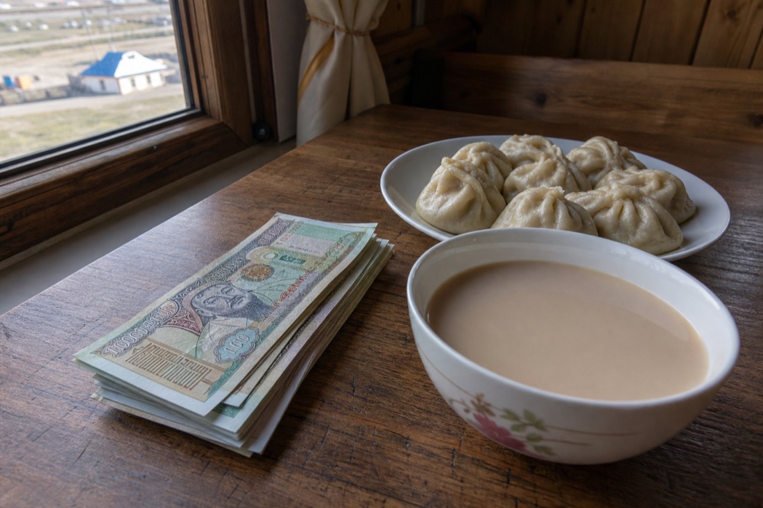 A stack of Mongolian tögrög banknotes beside a bowl of milk tea and dumplings on a wooden guesthouse table in Ulaanbaatar, morning light through the window