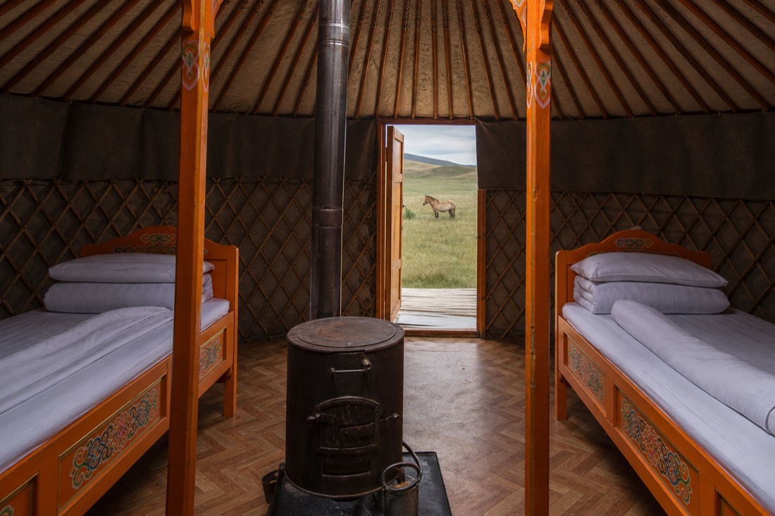 The interior of a traditional Mongolian guest ger at a Hustai tourist camp, with two painted orange wooden beds, a central iron stove with its chimney rising through the roof ring, and felt-lined lattice walls